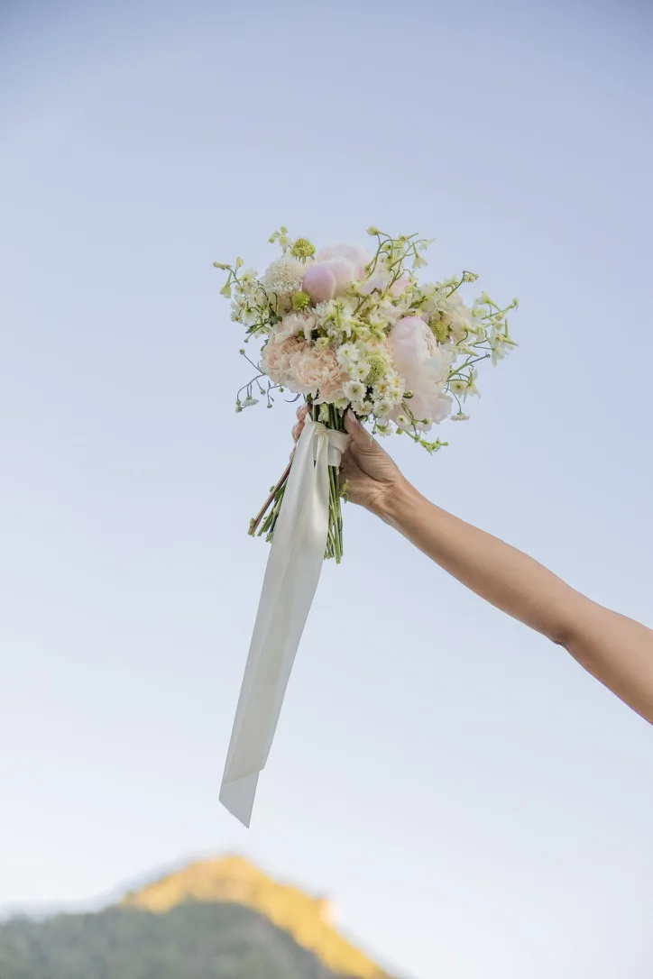A hand holds a soft white and green bouquet up against a clear pale sky, long ivory ribbons hanging from the stems.