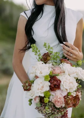 A bridal bouquet of blush peonies with deep burgundy blooms worked through it, held at the waist.