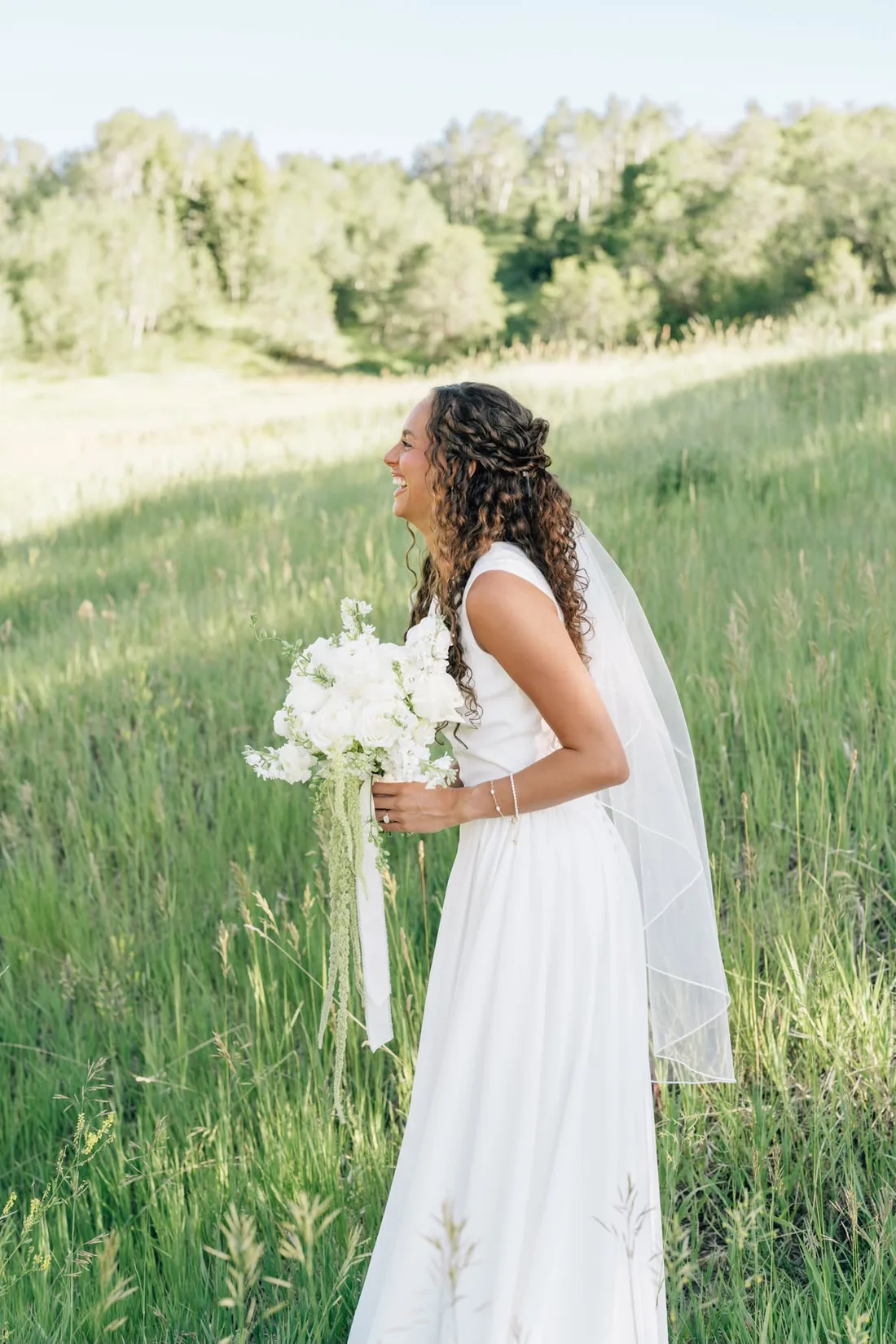 A bride in a long veil stands in tall summer grass holding an all-white bouquet with trailing greenery.