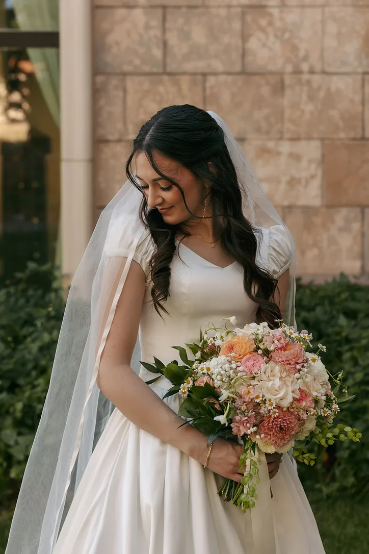 A bride in a veil looks down at a blush and peach bouquet, standing beside a stone building.
