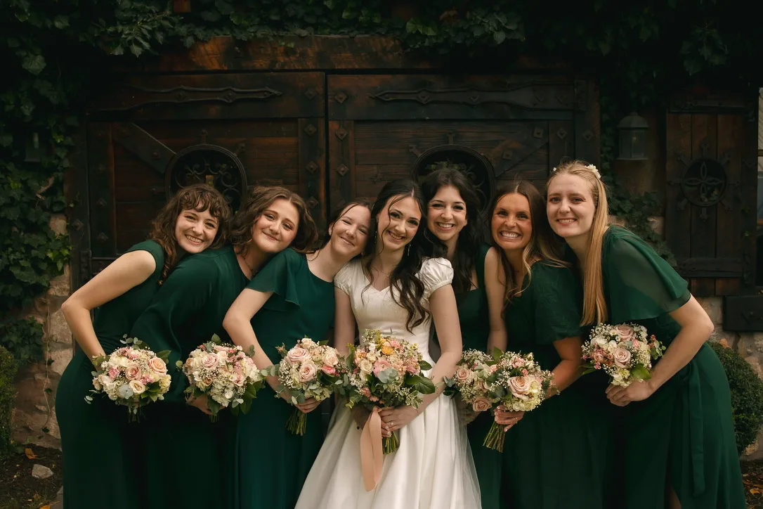 Six bridesmaids in dark green dresses stand together in front of a wooden door, each holding a blush and cream bouquet.