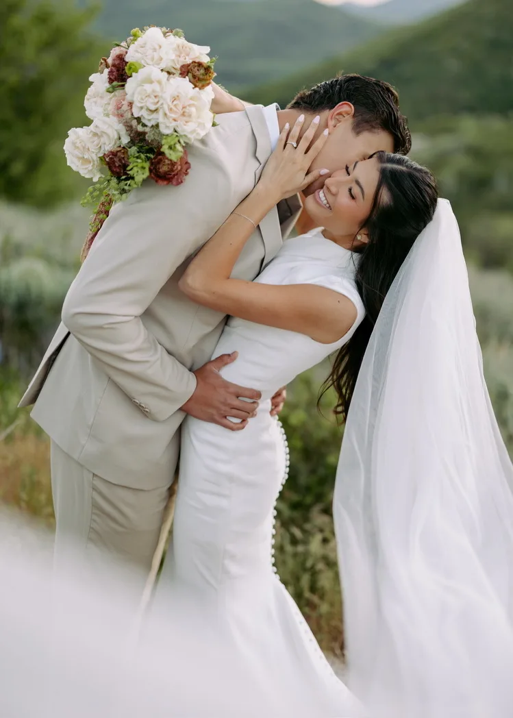 A groom lifts his bride off the ground in an embrace outdoors, her veil catching the light behind her.