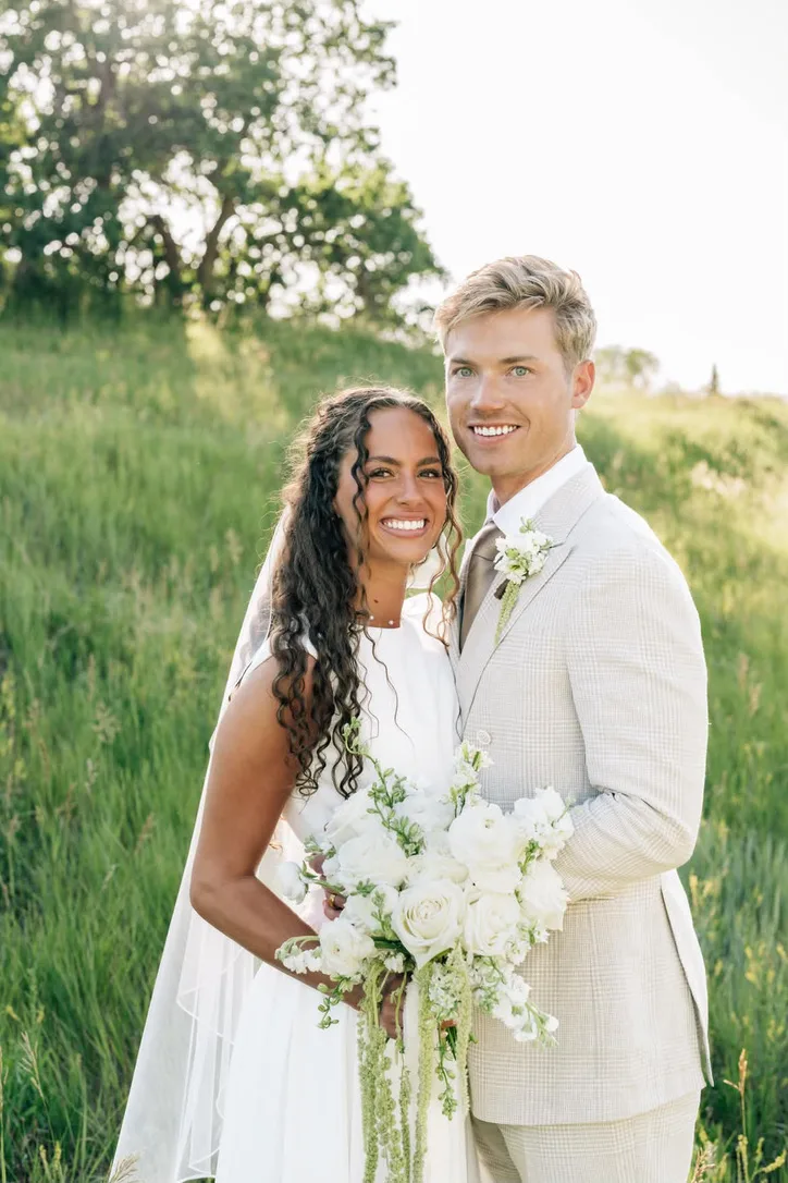 A couple stand together in a green field at golden hour, the bride holding a white and green bouquet.