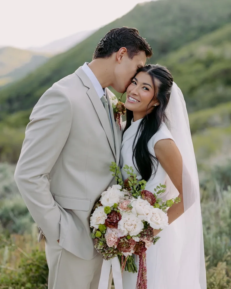 A couple stand cheek to cheek outdoors with a mountain slope behind them, the bride holding a blush and burgundy bouquet.