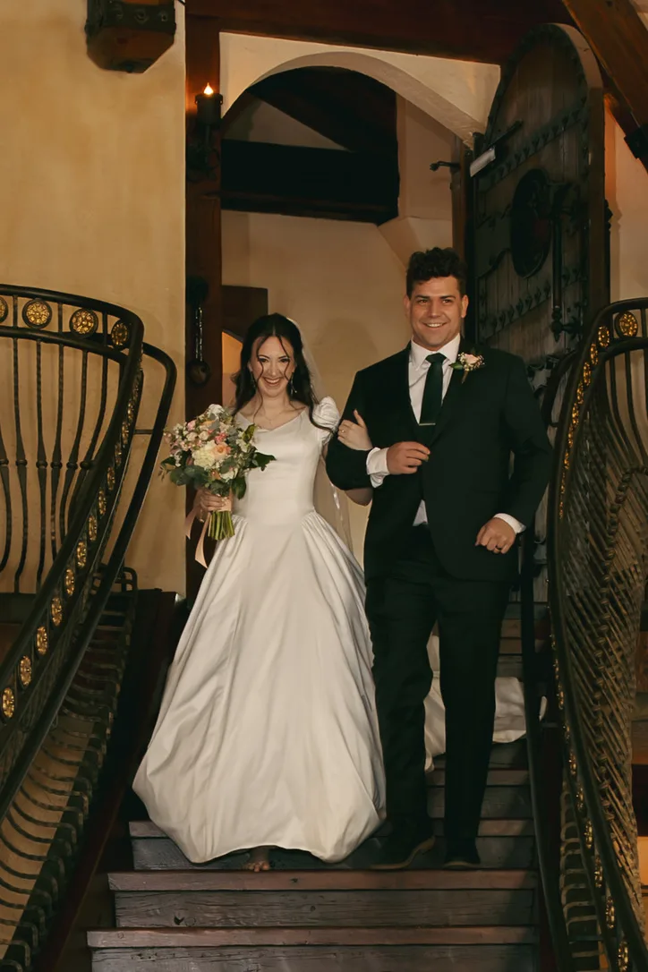 A couple descend an iron staircase arm in arm, the bride carrying a blush bouquet and the groom in a black tuxedo.