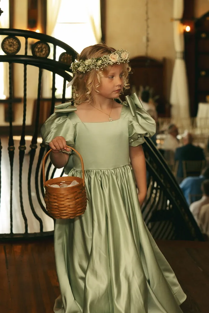 A flower girl in a sage green dress and a white flower crown carries a woven basket down a staircase inside a venue.