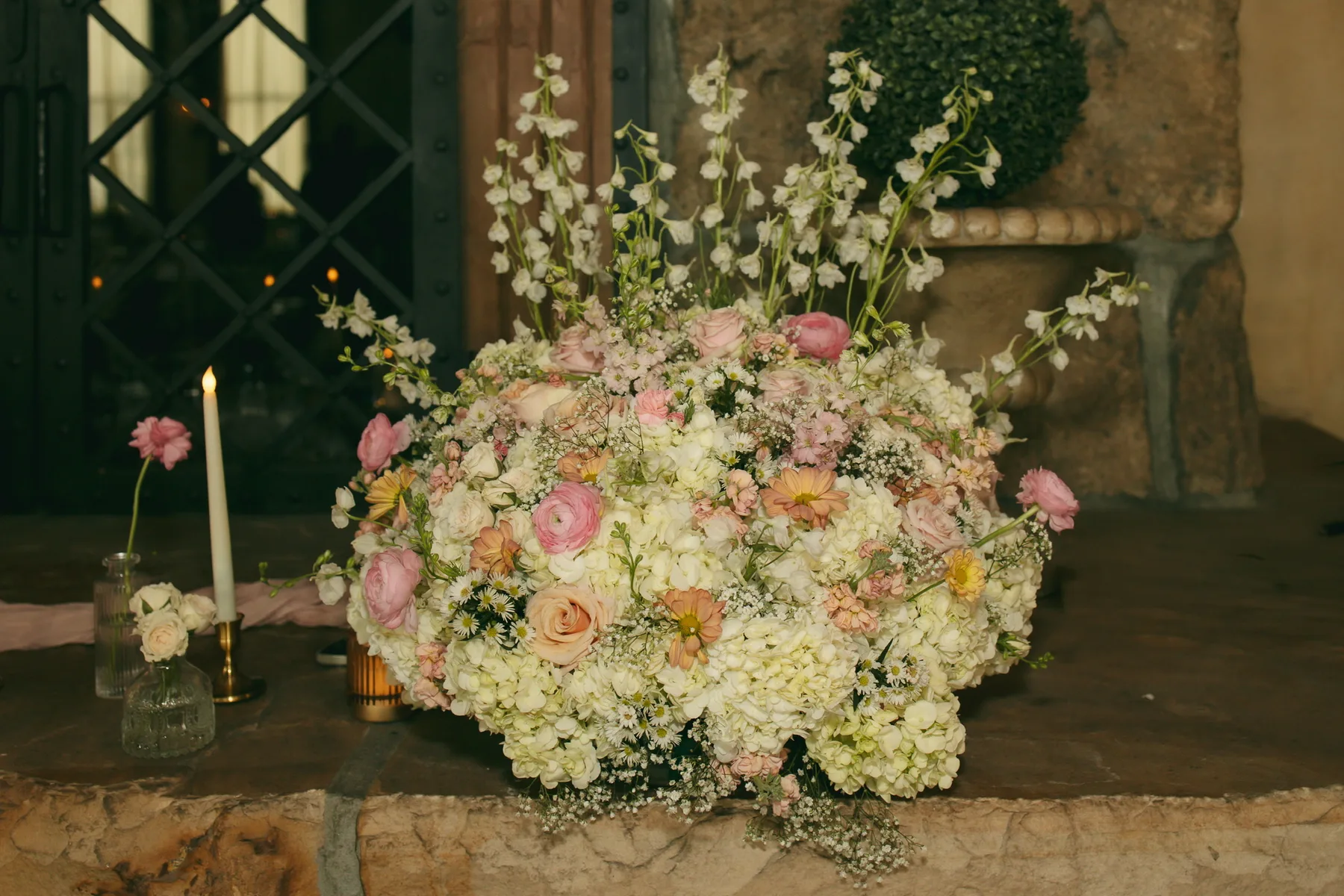 A full wedding centerpiece of pink and cream roses and white blossom spilling over its container, lit by tapered candles against dark greenery.