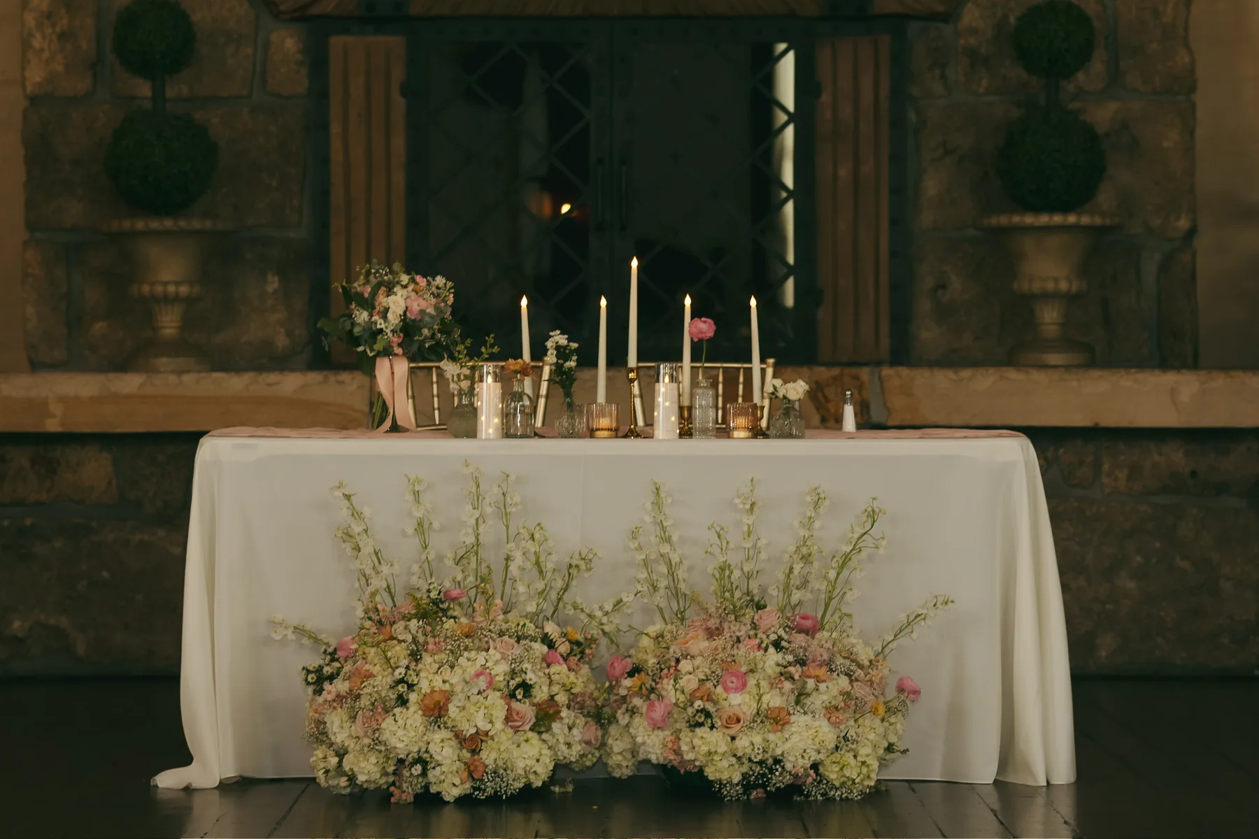 A long wedding head table runs the width of the frame under a garland of white and blush flowers and trailing greenery, with tapered candles in glass holders down its length.