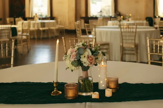 A round reception table with a dark green runner, amber glass votives and a small blush centerpiece.