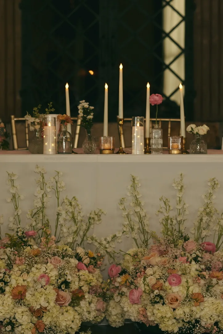 A long reception table dressed with tapered candles, glass votives and low sprays of greenery and pale blossom.